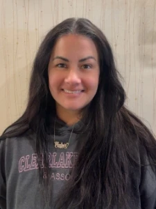Dental office team member with long dark hair smiling against wood panel wall.